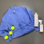Una giacca trapuntata blu è distesa su una superficie grigia, accompagnata da tre palline da tennis gialle e due flaconi di detersivo. La composizione mette in risalto i colori vivaci sullo sfondo neutro.