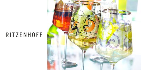 Un set di bicchieri Ritzenhoff colorati e artistici, pieni di bevande. Ogni bicchiere ha un design unico con motivi e figure vivaci. Lo sfondo mostra riflessi di luce e altri bicchieri, creando un'atmosfera festosa.