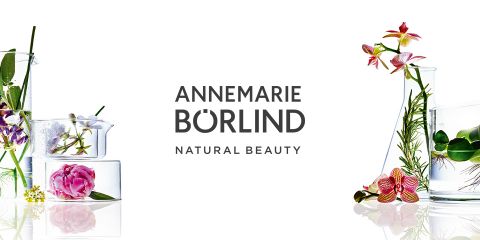 Contenitore di vetro con fiori ed erbe intorno, che mettono in risalto elementi naturali. Al centro c'è scritto "ANNEMARIE BÖRLIND NATURAL BEAUTY". Lo sfondo è bianco.