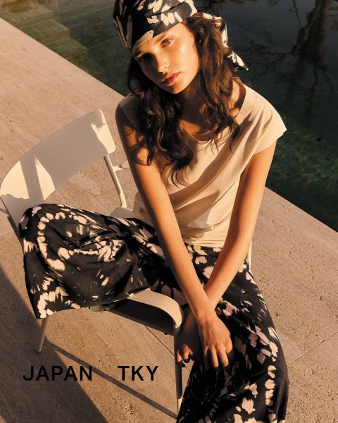 Una donna in top bianco e pantaloni a fiori è seduta su una sedia all'aperto con le braccia conserte. Indossa un foulard abbinato a fiori. In basso si legge il testo "JAPAN TKY". La scena è immersa in una calda luce solare.