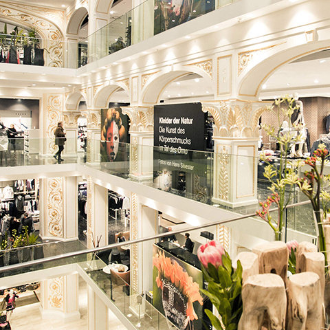 Un centro commerciale a più piani con interni decorati ad arte in bianco e oro. I clienti curiosano tra i negozi su diversi piani. Si notano grandi cartelloni pubblicitari e composizioni floreali. Le ringhiere in vetro e gli archi decorativi aggiungono eleganza alla scena.