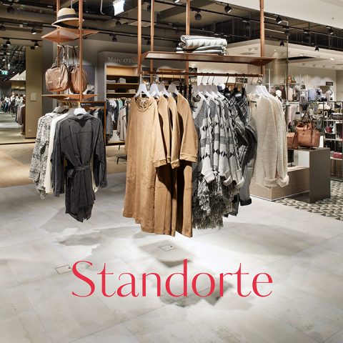 Un elegante interno di un negozio di abbigliamento con capi appesi in tonalità neutre come beige e grigio. Il negozio offre una varietà di articoli di moda e accessori. La parola "Standorte" è ben visibile sul pavimento.