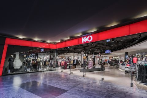L'immagine mostra l'esterno di un negozio di abbigliamento K&O in un centro commerciale. La facciata del negozio è in rosso e nero e presenta manichini che espongono vari outfit. All'interno si vedono appendiabiti accanto a luci luminose a soffitto.