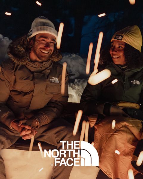 Due persone sedute attorno a un falò si sorridono. Indossano giacche e berretti di North Face. In primo piano si vedono scintille del fuoco. La scena è ambientata in un paesaggio notturno innevato con il logo di North Face sul bordo inferiore.