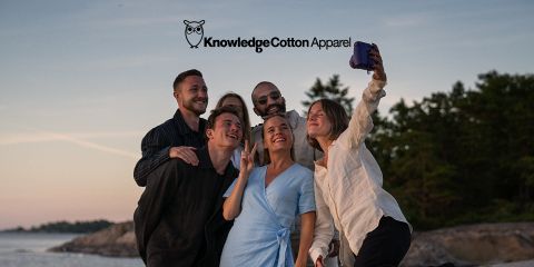 Un gruppo di cinque persone si fa un selfie all'aperto vicino all'acqua, con alberi e un tramonto sullo sfondo. Sorridono e posano in modo giocoso, vestiti in abiti casual. Sopra di loro si vedono il testo "KnowledgeCotton Apparel" e un logo a forma di gufo.