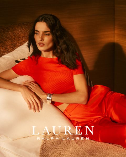 Una donna con lunghi capelli ondulati è appoggiata su un divano bianco, indossa un top rosso brillante e pantaloni abbinati. La calda luce del sole proietta ombre sulla scena. In basso si legge "LAUREN RALPH LAUREN".