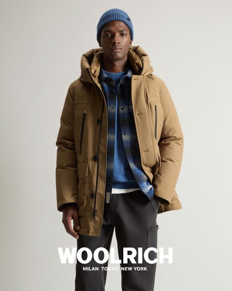 Una persona che indossa un cappotto invernale marrone chiaro, un berretto di lana blu e una camicia a quadri blu sopra una felpa con cappuccio, si trova di fronte a uno sfondo a tinta unita. Sotto appare la parola "Woolrich" insieme alle città di Milano, Tokyo e New York.
