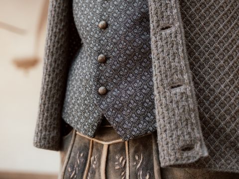 Primo piano di una persona che indossa un cardigan grigio lavorato a maglia sopra un gilet grigio fantasia con tre bottoni rotondi. La parte inferiore mostra una gonna con pieghe e delicati dettagli ricamati. Lo sfondo è sfocato.
