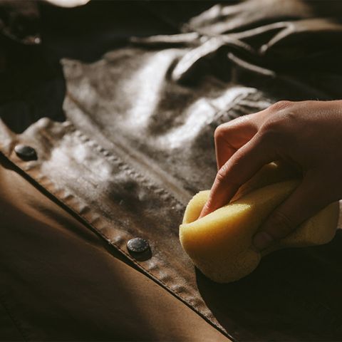 Una mano pulisce o lucida una giacca di pelle marrone con una spugna gialla, concentrandosi su bottoni e cuciture. L'illuminazione mette in risalto la struttura e la lucentezza della pelle.