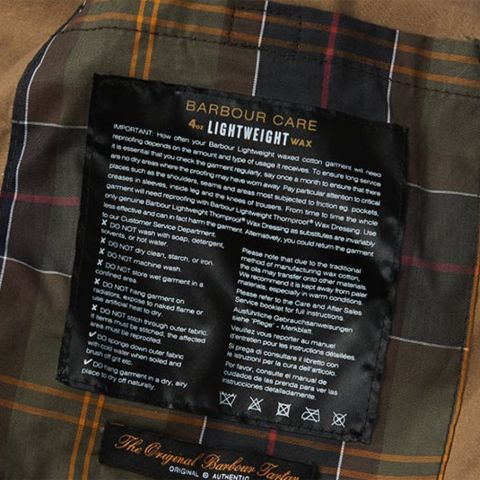Primo piano di un'etichetta di manutenzione su tessuto scozzese. L'etichetta contiene istruzioni per la cura dell'abbigliamento Lightweight Wax di Barbour, nonché consigli per la pulizia e la manutenzione. Lo sfondo mostra un motivo scozzese marrone, verde e arancione.