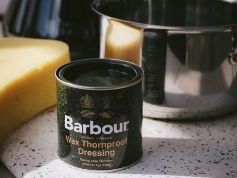 Una latta di Barbour Wax Thornproof Dressing è appoggiata su una superficie screziata accanto a una grande spugna gialla e un lucido pentolino di metallo. La latta è scura con scritte bianche e dorate e viene utilizzata per impermeabilizzare gli indumenti.