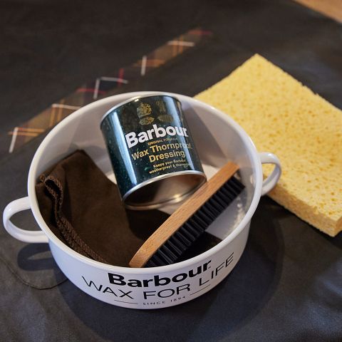 Un kit di manutenzione Barbour con una lattina di cera, un panno per lucidare, una spazzola e una spugna gialla. Gli articoli sono disposti in un contenitore rotondo bianco con il testo "Barbour Wax for Life".