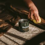 Una persona applica cera su un indumento marrone con una spugna gialla. Un barattolo con la scritta "Barbour Wax Thornproof Dressing" è posizionato nelle vicinanze su una superficie rivestita di tessuto. La scena è illuminata da una luce calda e le ombre suggeriscono una fonte di luce naturale.