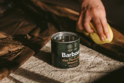 Una persona applica cera su un indumento marrone con una spugna gialla. Un barattolo con la scritta "Barbour Wax Thornproof Dressing" è posizionato nelle vicinanze su una superficie rivestita di tessuto. La scena è illuminata da una luce calda e le ombre suggeriscono una fonte di luce naturale.