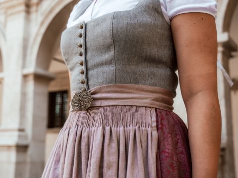 Primo piano di una persona in abiti tradizionali con un corpetto grigio aderente, bottoni decorati e un'ampia gonna nei toni del lavanda e del rosso. Si trova di fronte a uno sfondo di archi in pietra.