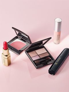 Una serie di prodotti per il trucco, tra cui rossetto, fard, palette di ombretti, mascara e fondotinta, disposti su una superficie rosa.