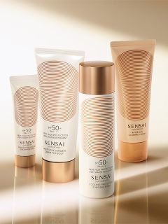 Quattro prodotti solari Sensai con packaging bianco e oro sono presentati su una superficie riflettente con uno sfondo caldo e luminoso.
