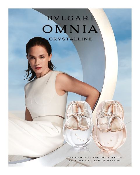 Una donna in abito bianco si appoggia sullo sfondo di un'architettura moderna sotto un cielo blu. In primo piano, due flaconi del profumo Bvlgari Omnia Crystalline, che presentano tonalità chiare e ambrate.