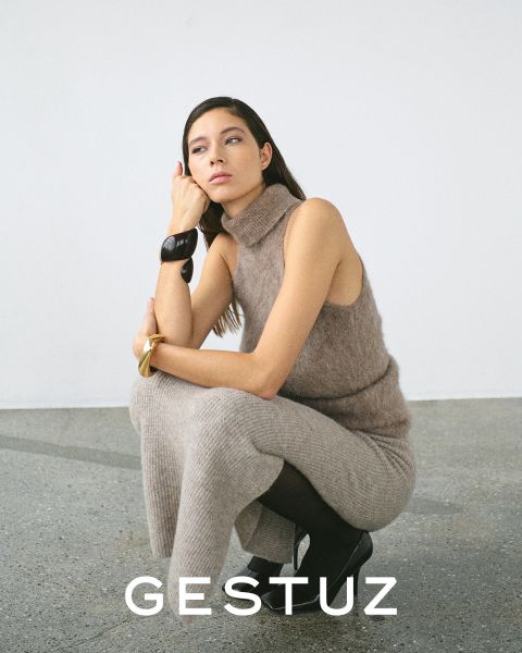 Una donna posa in un accogliente dolcevita senza maniche in maglia beige e pantaloni abbinati. È accovacciata con un ginocchio sollevato, indossa stivali neri e un braccialetto grosso. Lo sfondo è minimalista. In basso c'è scritto "GESTUZ".