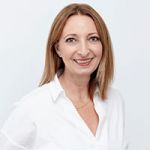 Una donna con capelli castano chiaro di media lunghezza sorride alla fotocamera. Indossa una camicetta bianca e una collana dorata e si trova di fronte a uno sfondo chiaro e semplice.