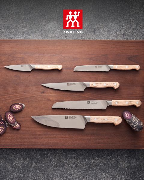 Cinque coltelli da cucina con manici in legno chiaro sono appoggiati su una base di legno, accanto ci sono carote viola tagliate a fette, e sopra è visibile il logo Zwilling.