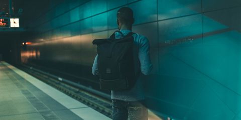 Una persona con uno zaino è in piedi su una banchina della metropolitana vuota e guarda i binari. La stazione è illuminata fiocamente da una luce blu-verde, creando un'atmosfera suggestiva. Un orologio sul soffitto indica l'ora, non si vede nessun treno.