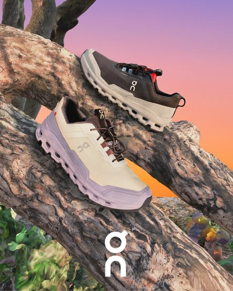 Due sneaker del marchio "On" sono presentate su un ramo strutturato di fronte a un cielo colorato al tramonto. Una scarpa è nera con accenti grigi e l'altra è bianca con dettagli più scuri. Sotto è visibile il logo "On".