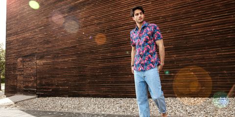 Una persona è in piedi all'aperto e indossa una camicia colorata con motivo floreale e jeans azzurri. Si trova di fronte a una parete di legno, sotto la quale c'è un sentiero di ghiaia. La luce del sole crea riflessi dell'obiettivo nell'immagine.