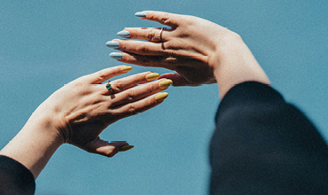 Due mani con unghie colorate, una in azzurro pastello e l'altra in giallo brillante, si stagliano contro un cielo azzurro limpido. Le mani indossano diversi anelli e lo sfondo appare morbido e sfocato.