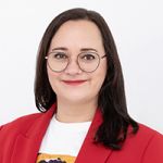 Una persona sorridente con lunghi capelli scuri e occhiali, che indossa un blazer rosso sopra una camicia bianca, si trova di fronte a uno sfondo bianco semplice.
