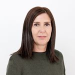 Una donna con capelli castani di media lunghezza e un maglione verde scuro è in piedi davanti a uno sfondo bianco semplice. Guarda la fotocamera con un'espressione neutra.