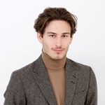 Una persona con capelli castani e baffi indossa un blazer grigio sopra un dolcevita beige. Sorride leggermente e guarda la fotocamera su uno sfondo bianco semplice.