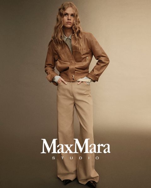 Una persona con i capelli lunghi indossa una giacca di pelle marrone sopra una camicia chiara e pantaloni beige a gamba larga. Si trova di fronte a uno sfondo beige sfumato. In basso si legge il testo "MaxMara Studio".