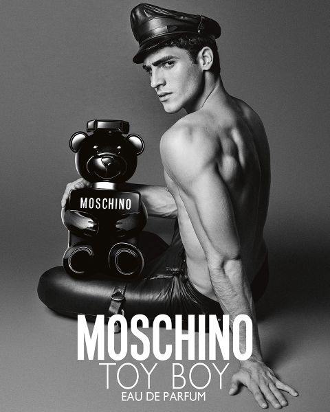 Uomo a torso nudo in lederhosen e cappello tiene in mano una bottiglia di profumo Moschino a forma di orsacchiotto. Testo: "MOSCHINO TOY BOY EAU DE PARFUM."