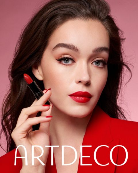 Una donna con la pelle chiara e i capelli scuri posa davanti a uno sfondo rosa, tiene in mano un rossetto rosso e indossa un blazer rosso e un rossetto rosso abbinato. Sotto c'è la scritta "ARTDECO".