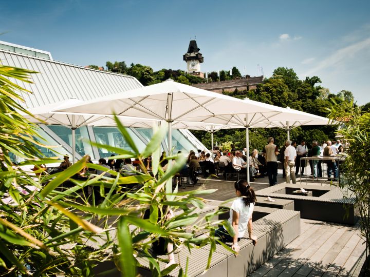 Persone si godono una giornata di sole in un caffè all'aperto con grandi ombrelloni. Sullo sfondo, un campanile storico si erge su una collina verde. Fogliame lussureggiante e architettura moderna circondano l'area salotto.