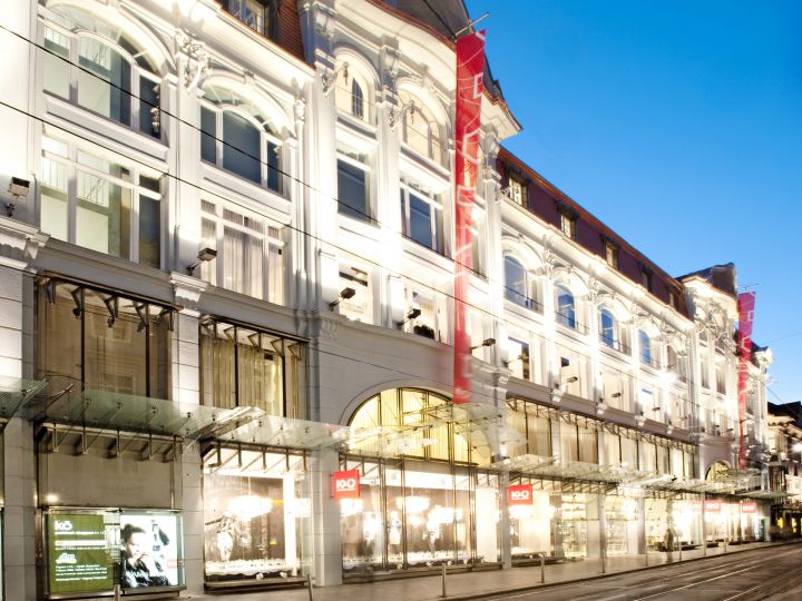Un grande magazzino illuminato con una facciata sfarzosa, molte finestre ad arco e striscioni rossi. La strada antistante è tranquilla e il cielo è limpido e blu, suggerendo il crepuscolo.