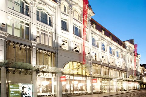Facciata illuminata di un grande ed elegante centro commerciale di sera, con finestre ad arco e tetti di vetro. La strada è vuota e le vetrine sono illuminate a giorno.