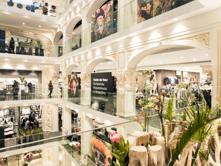 Un elegante centro commerciale a più piani con dettagli architettonici classici. I clienti possono curiosare tra i negozi con ampie vetrine alla ricerca di abbigliamento. Piante e fiori decorativi sono disposti attorno all'atrio aperto.