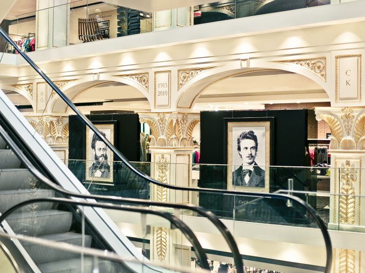 Interno di un grande magazzino decorato in modo elaborato con scale mobili e due ritratti incorniciati di un uomo esposti al piano superiore. Lo spazio è caratterizzato da colonne decorative e illuminazione elegante.
