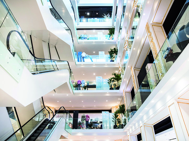 Interno di un moderno centro commerciale a più piani con illuminazione brillante, ringhiere in vetro, scale mobili e aree verdi su ogni piano. Il design aperto presenta diversi piani e vetrine dei negozi in un ambiente spazioso.