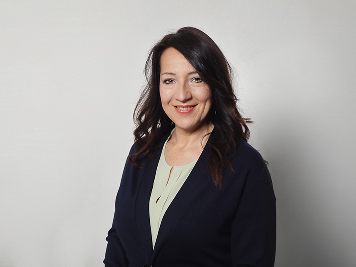 Una donna con lunghi capelli scuri, un blazer blu navy e una blusa chiara sorride alla telecamera su uno sfondo bianco semplice.