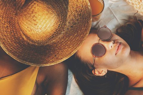Due persone si rilassano su una coperta da spiaggia. Una indossa un grande cappello di paglia che le copre il viso, l'altra indossa occhiali da sole e le sta accanto. Entrambi indossano costumi da bagno e hanno un bicchiere di succo d'arancia vicino, creando un'atmosfera estiva e rilassata.