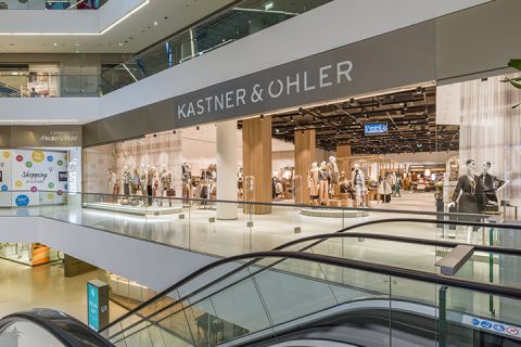 Interno di un moderno centro commerciale con il negozio Kastner & Öhler. Sulla facciata del negozio si vedono manichini con vari outfit. In primo piano si vedono le scale mobili e il centro commerciale ha diversi piani con ringhiere in vetro.
