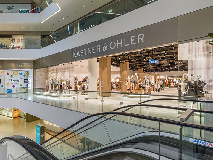 Interno di un moderno centro commerciale con il negozio Kastner & Öhler. Sulla facciata del negozio si vedono manichini con vari outfit. In primo piano si vedono le scale mobili e il centro commerciale ha diversi piani con ringhiere in vetro.
