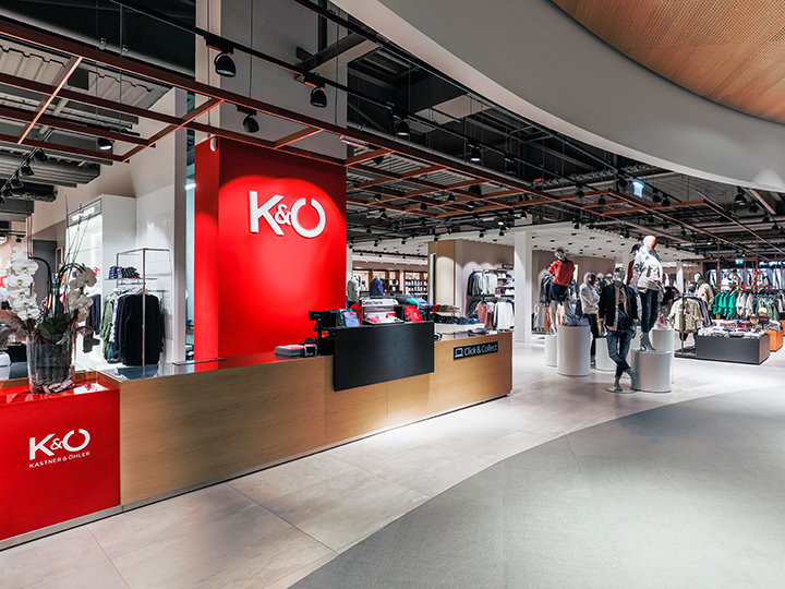 Interno di un moderno negozio di abbigliamento con un bancone rosso acceso che mostra il logo K&Ö. Nel negozio si vedono diversi appendiabiti con vestiti, sullo sfondo manichini e una persona che curiosa. L'atmosfera è spaziosa e ben illuminata.