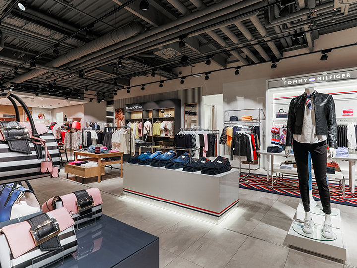 Un interno moderno di un negozio di abbigliamento con vari capi. In primo piano, manichini presentano outfit alla moda. Gli scaffali espongono indumenti piegati e sui tavoli sono esposti accessori. Un'illuminazione brillante mette in risalto il design moderno del negozio.