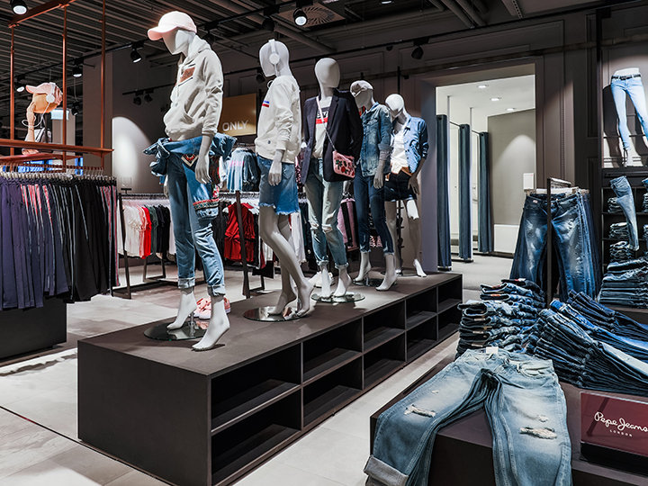 In un negozio di abbigliamento, dei manichini espongono abiti casual in jeans, tra cui jeans e giacche. Gli scaffali circostanti contengono vari capi di abbigliamento e su uno scaffale si può vedere una pila di jeans piegati. Il negozio ha un design moderno.