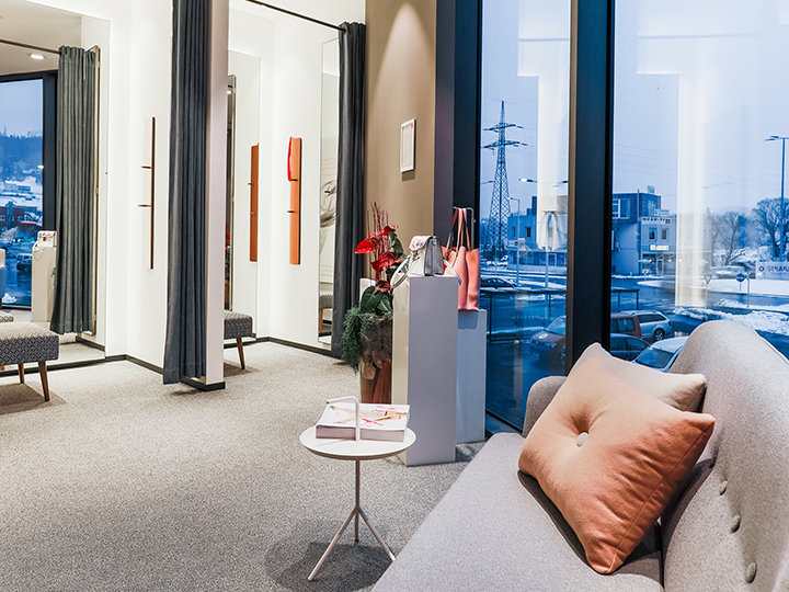 Una moderna ed elegante sala d'attesa con un divano, un tavolino rotondo e ampie finestre che offrono una vista su un paesaggio urbano innevato. La stanza è dotata di moquette, tende e piante decorative su un piedistallo.