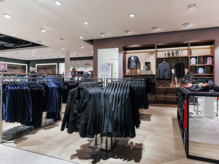 Un moderno negozio di abbigliamento con appendiabiti pieni di abiti appesi ordinatamente. Sugli scaffali sullo sfondo si possono vedere camicie piegate, blazer e accessori. Il negozio è ben illuminato e ha un layout elegante e ordinato.